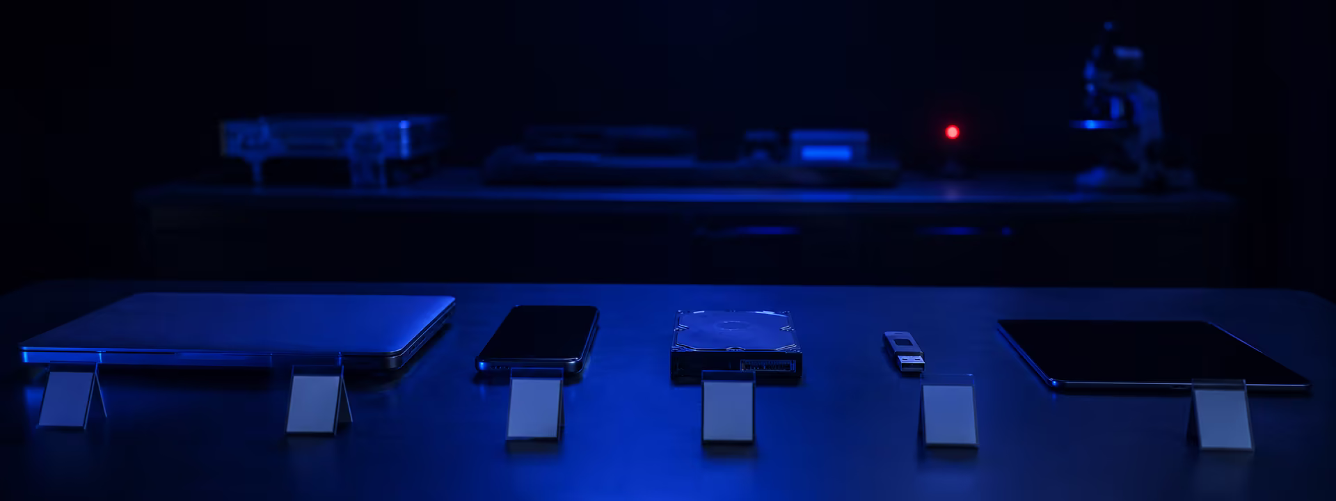 Laptop, teléfono, disco duro, memoria USB y tableta alineados y etiquetados sobre una mesa de laboratorio